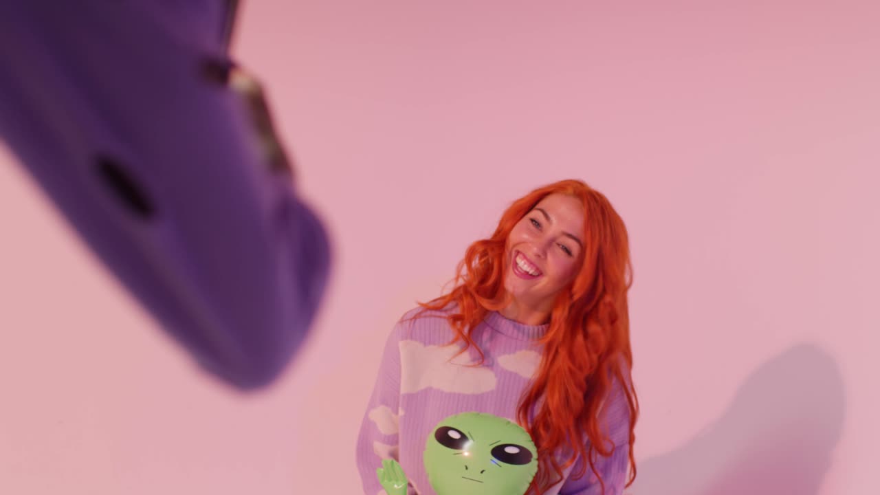 scatto in studio di una donna che scatta una foto di un'amica che gioca con un alieno gonfiabile su un telefono cellulare sullo sfondo rosa