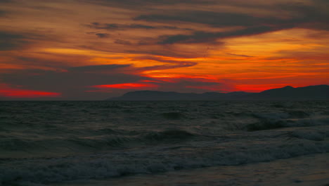 4k uhd cinemagraph bucle de vídeo sin fisuras de la puesta de sol vista desde una romántica playa de arena con montañas y olas en la hermosa costa mediterránea italiana con nubes rojas y naranjas en el cielo