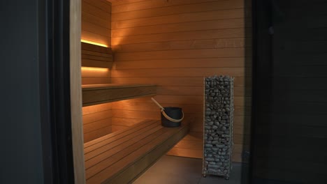 slow push inside of a luxury wooden cedar wood sauna complete with tall rock heat, 2 level benches and floor to ceiling window showing view into forested area