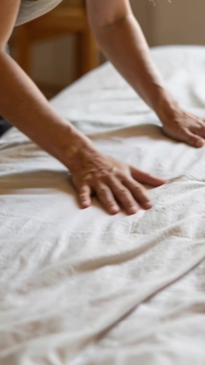 Video vertical: Mujer con top claro alisando una sábana en la cama del dormitorio, dejando la superficie plana.