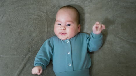adorable baby lying on the blanket and looking into camera. calm baby boy in blue suit at grey background. view from above.