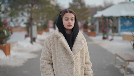 thoughtful lady walking outdoors in warm beige winter coat and black hoodie on cold snowy day, background features snow-covered ground, dry trees, and festive decorations in soft blur