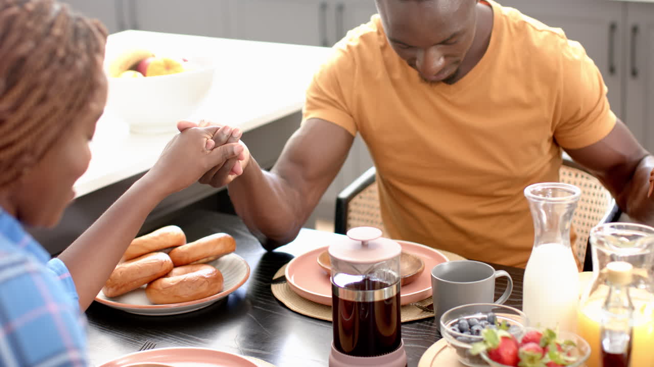 african american family holding hands and praying before lunch, with food on table