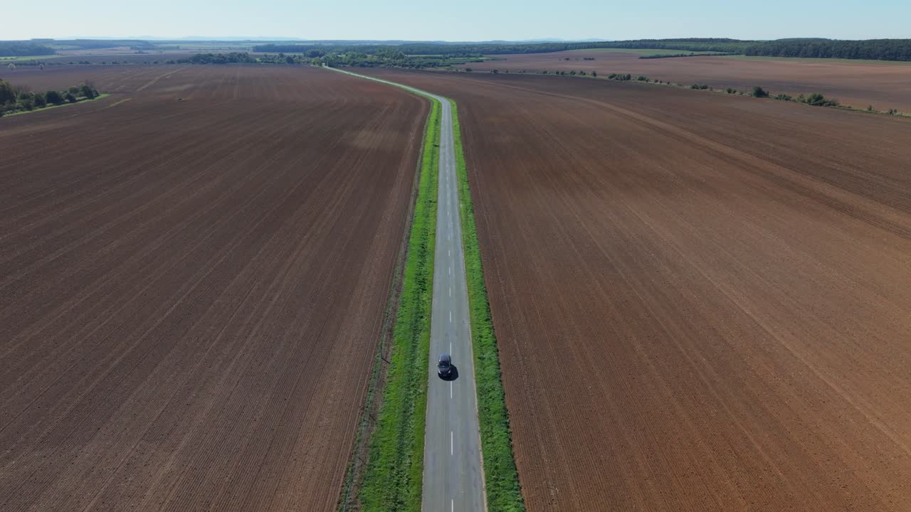 An aerial view of a long straight country road cutting through vast plowed fields. The landscape stretches to the horizon with farmland on both sides, showcasing rural simplicity and open space