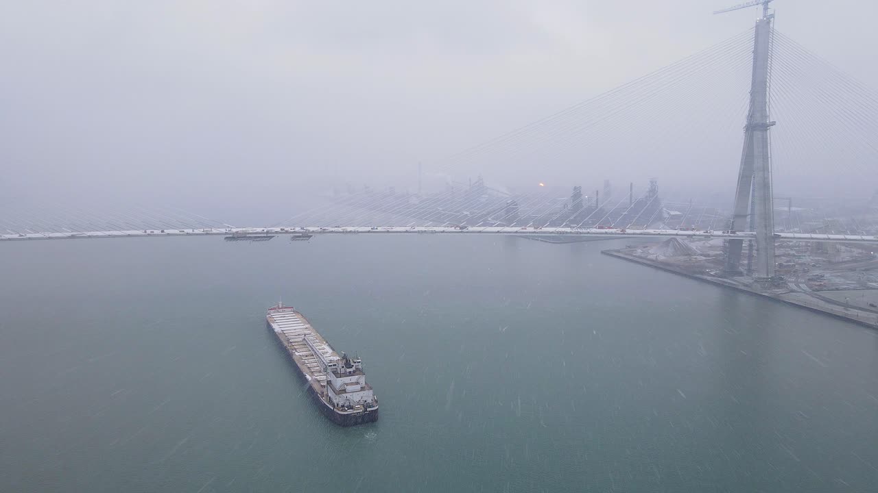 Cargo ship passes beneath Gordie Howe Bridge near snowy industrial banks of Detroit River