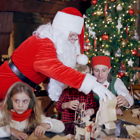 cheerful children are playing with wooden toys with the help of elves and santa claus. two elves in green costumes, santa and little children on the background of christmas tree at home.