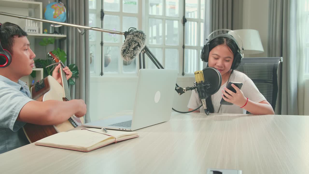 Asian Kid Girl Holding Phone And Wearing Headphones Singing Into Microphone While Boy Playing The Guitar During Online Livestream In Podcast Studio