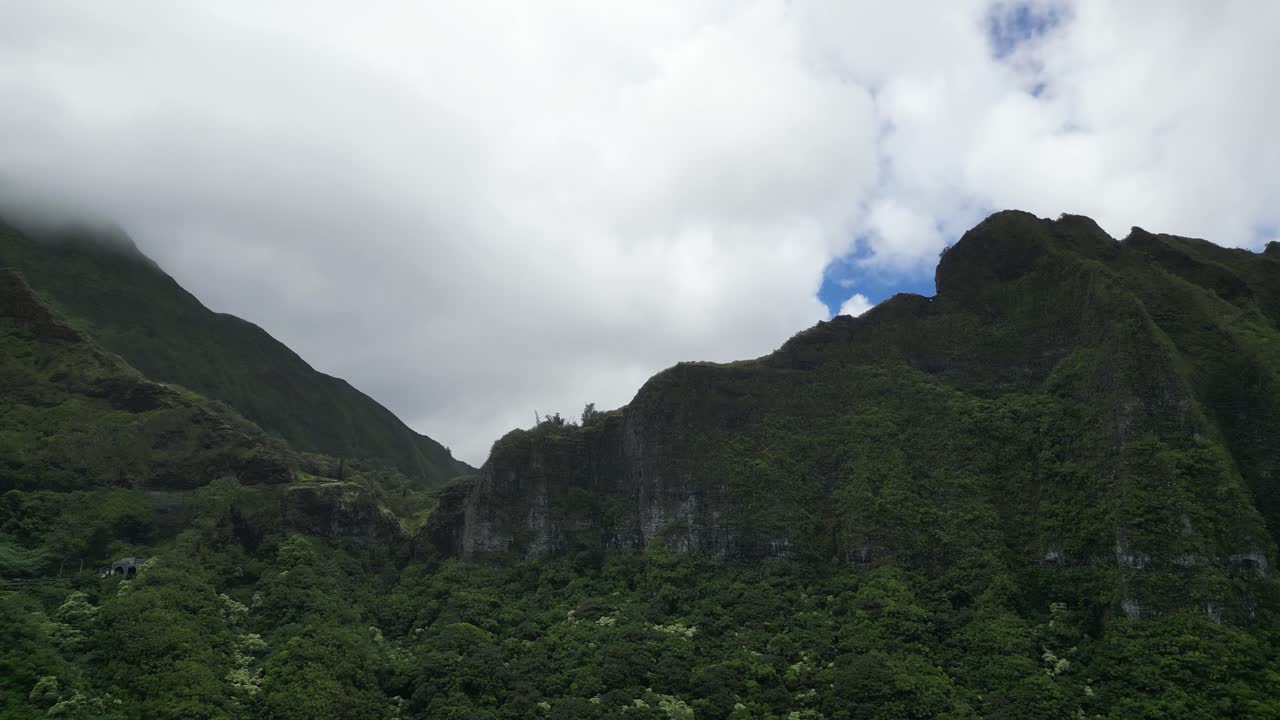 Drone captures Hawaii’s forested mountains from below with cloudy sky and steep cliffs.