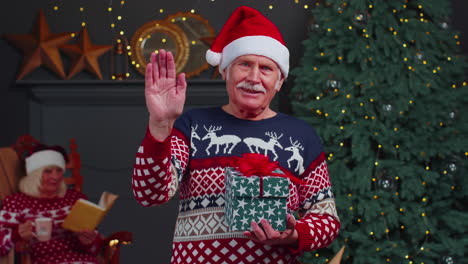 Senior-grandfather-in-Christmas-sweater-smiling-friendly-at-camera-and-waving-hands-gesturing-hello