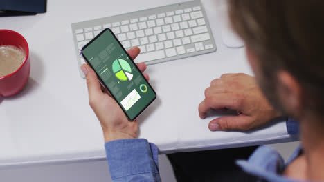 hombre caucásico sentado en un escritorio usando un teléfono inteligente con estadísticas en la pantalla