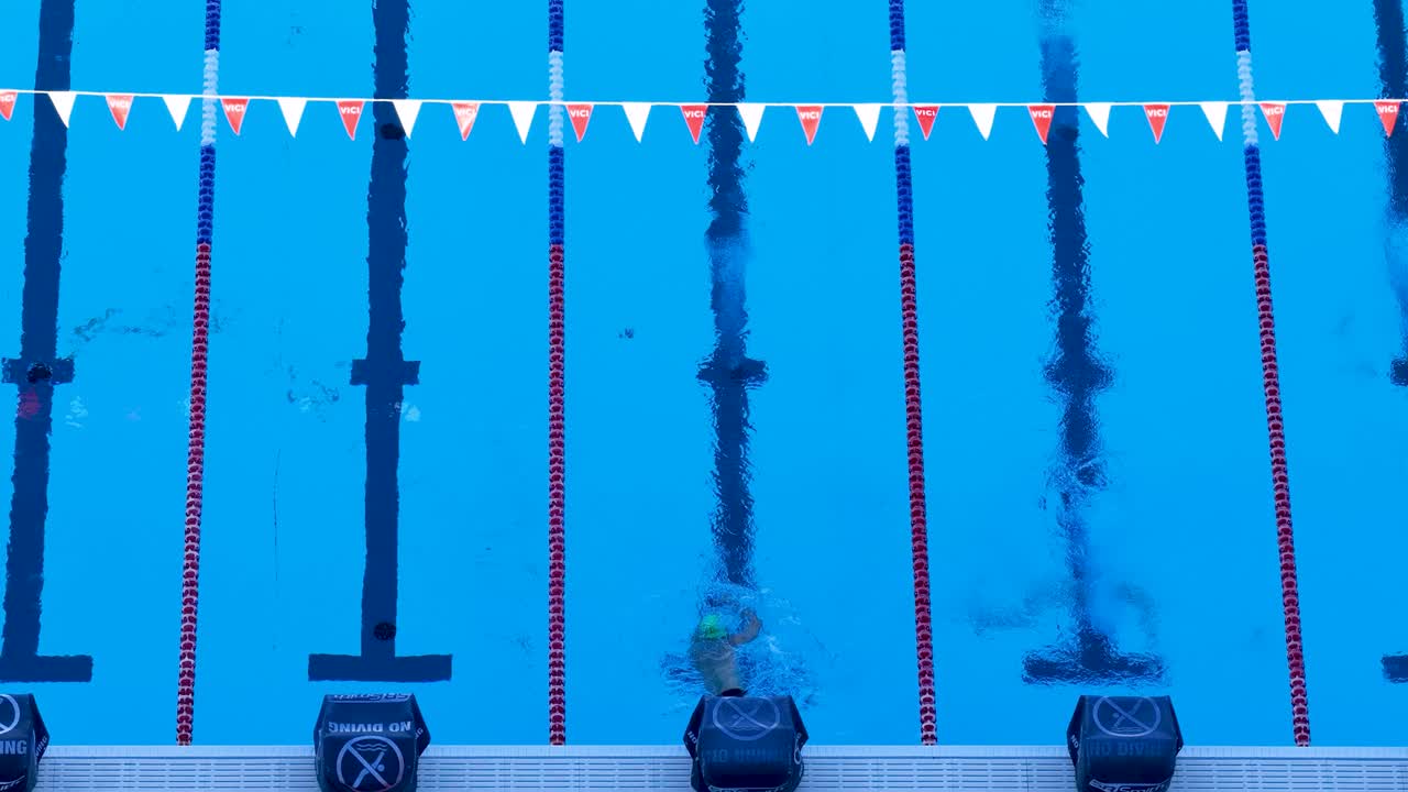 Drone captures swimmer practicing freestyle in a clear blue pool. Bright lighting and steady camera enhance the serene aquatic scene