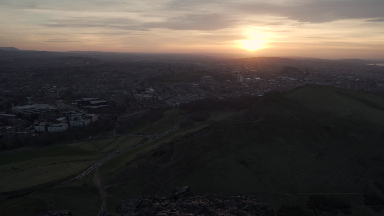 toma panorámica del paisaje urbano de edimburgo desde la montaña arturs seat durante la puesta de sol con colores maravillosos y luz dorada con ulex bush en primer plano y autos que pasan por debajo