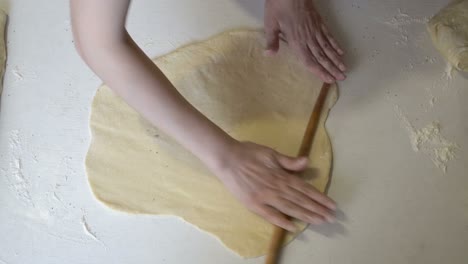 a female chef rolls out a thin layer of dough with a wooden rolling pin in the kitchen, on a white table. the concept of cooking. close-up. the view from the top. faceless. 4k.