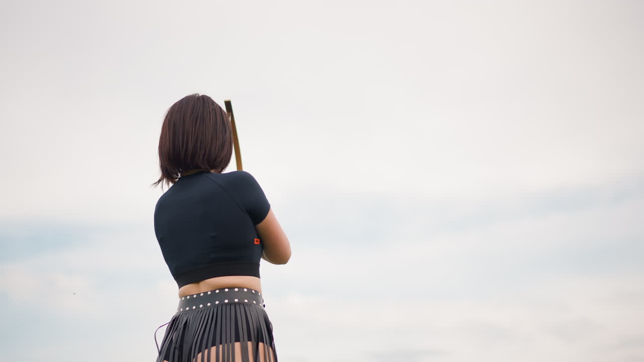black woman archer facing sky, windswept horizon, steady stance holding traditional bow, contemplative mood, soft clouded sky, solitary warrior presence, focused gaze toward distant target, subtle