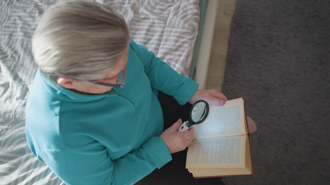 Senior woman reading book with magnifying glass in hand. Close-up of elderly woman scanning text using magnifier, focused on detailed reading, soft natural light illuminating her hands and book