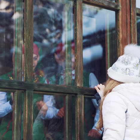 a little girl with a letter in her hands is standing on the street and looking out of the window behind the elves with long ears in green suits who are dancing.