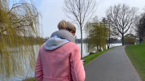 Tracking-shot-of-fashionable-young-girl-checking-mobile-phone-during-walk-on-bank-of-river-during-beautiful-day