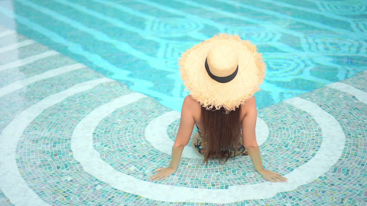 Rückansicht einer hübschen Frau im tropischen Spa, am Pool sitzend, mit Hut und Badeanzug, Draufsicht