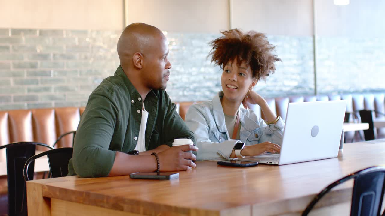 African couple sitting at cafe table, reviewing laptop screen, discussing work, man holding coffee