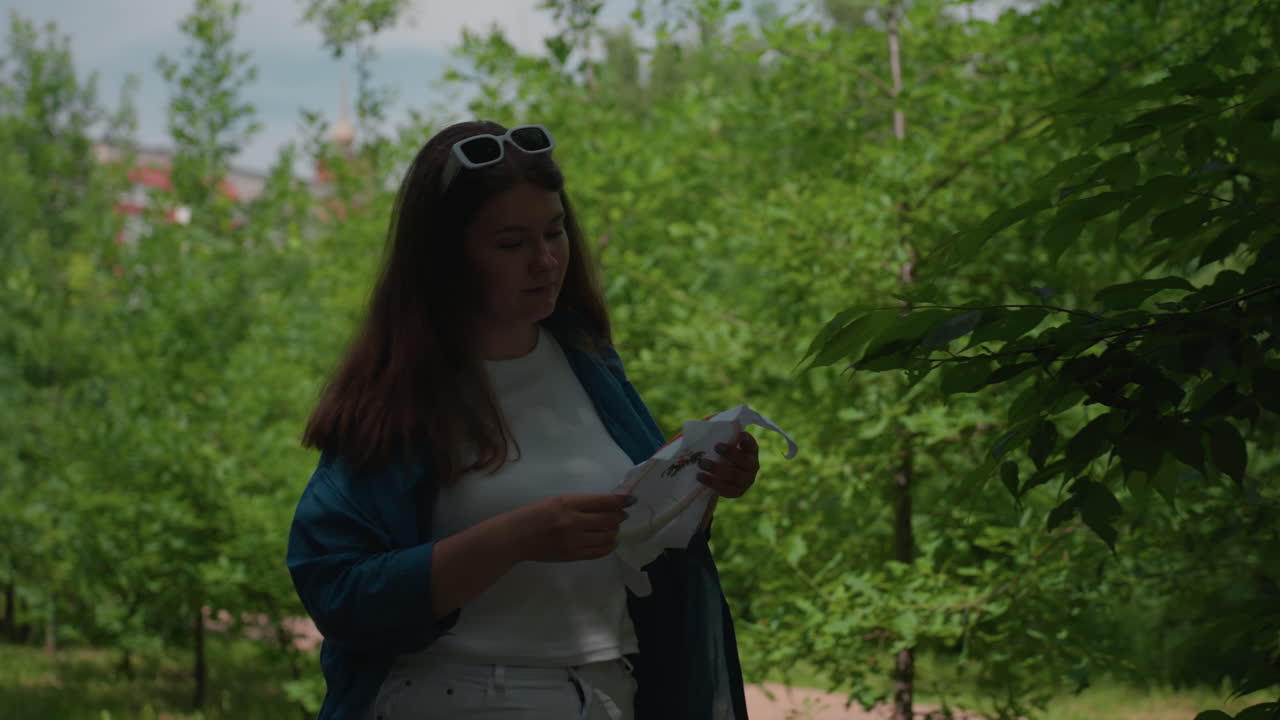 Young girl in white outfit and blue shirt strolling along lush green alley holding embroidered fabric, admiring her handmade work with pride while dancing lightly