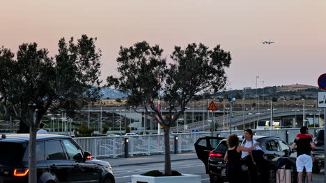 evening scene at an airport with cars, people, and a plane in the sky