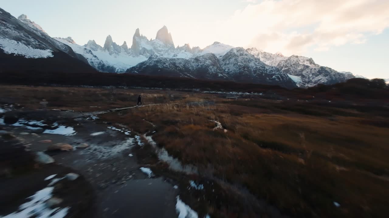Stunning FPV aerial of Fitz Roy mountain, Patagonia, flying past a lone hiker on a trail toward the vast, beautiful open landscape. Snowy terrain glowing in sunlight.
