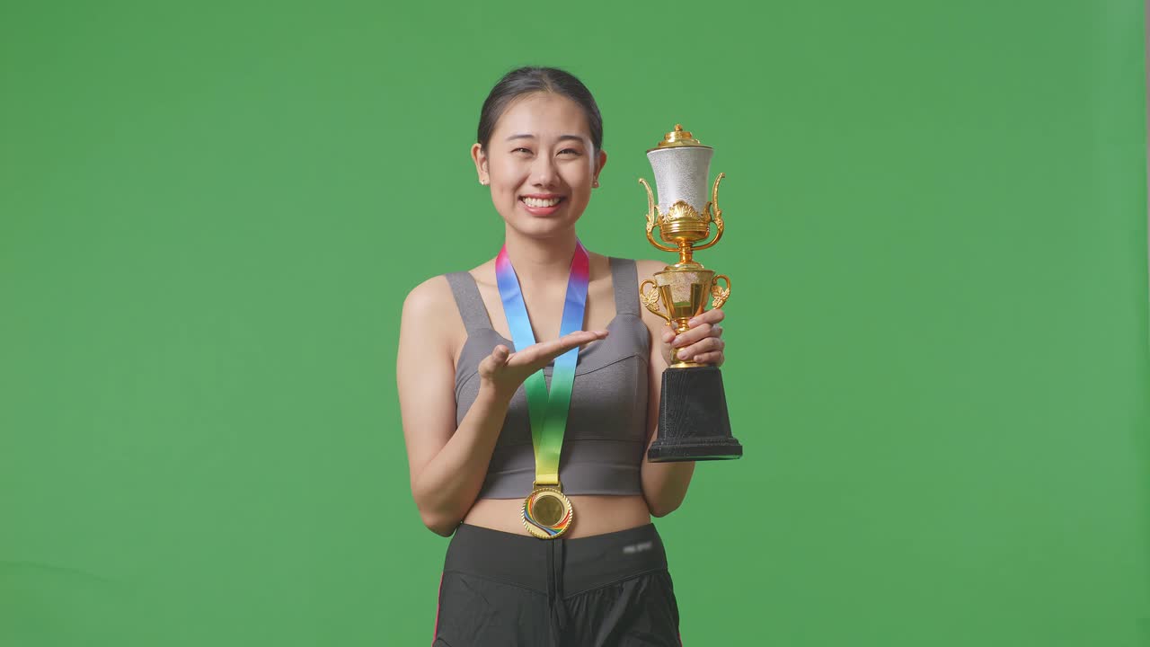 금메달을  아시아 여성은 손에 금 트로피를 가리키며 미소 짓고 스튜디오의 녹색 화면 배경에서 첫 번째 우승자로 자랑스럽게 승리합니다.