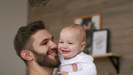 Loving-father-in-white-t-shirt-hugs-the-baby-and-kisses-the-baby-laughs-and-smiles-on-the-background-of-the-Christmas-tree-and-garlands