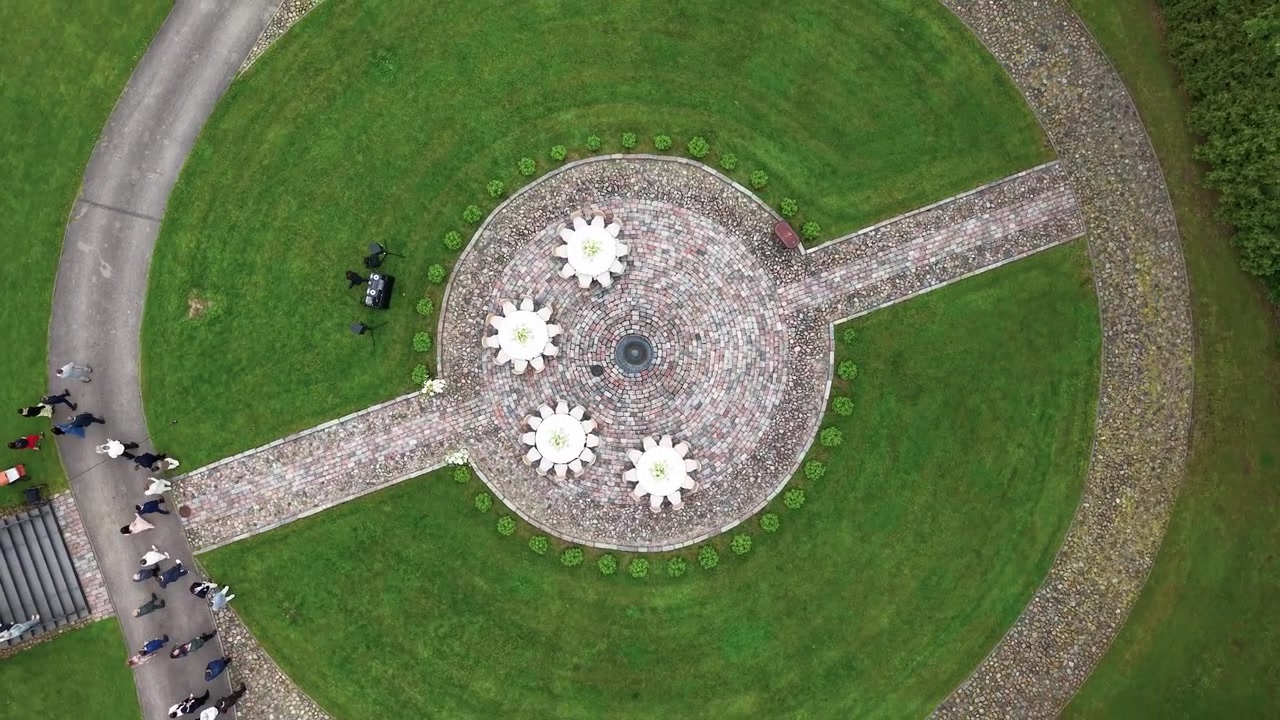 Vídeo de drone captura pessoas chegando por caminhos de pedra bem cuidados a um pátio circular com decorações em forma de flor, cercado por um gramado verde exuberante e paisagismo formal, sob luz nublada