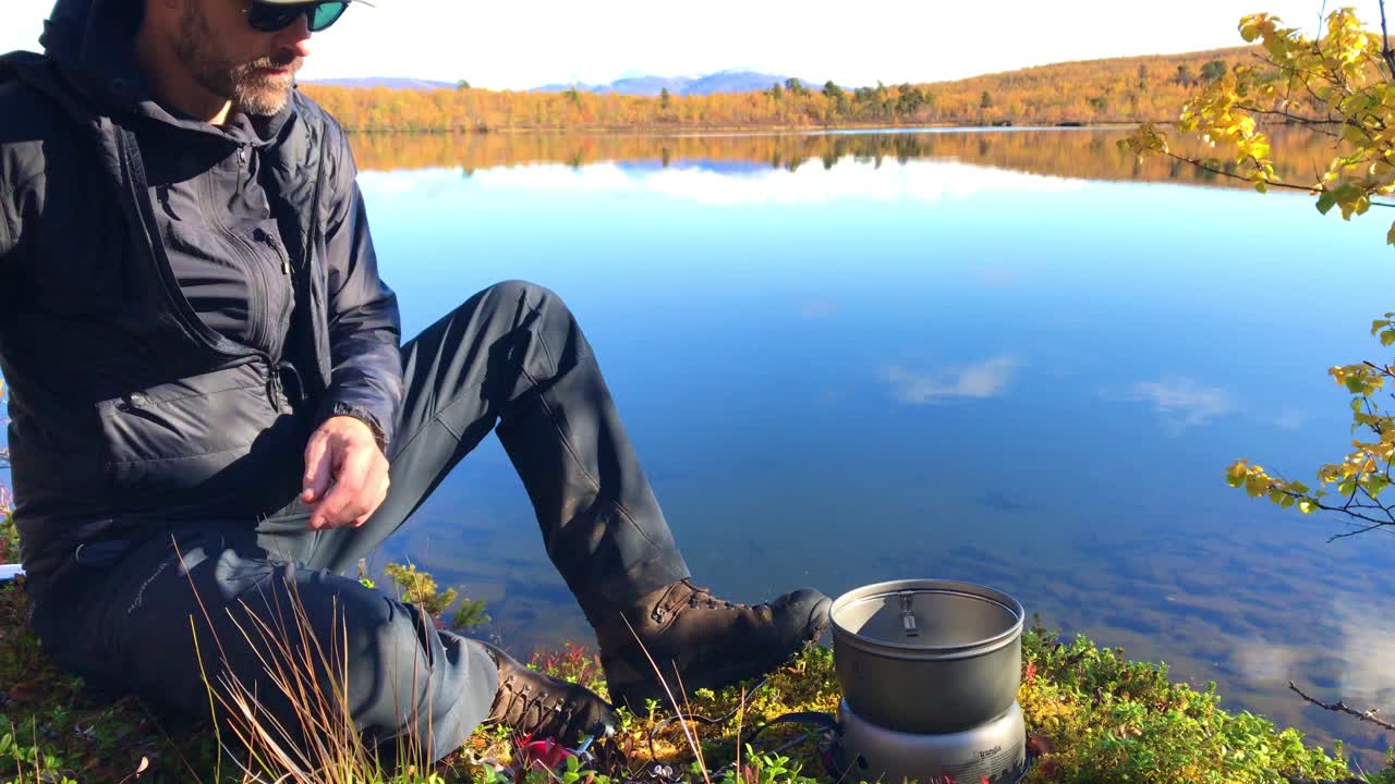 Un escursionista seduto accanto a un lago di montagna autunnale incredibilmente limpido e calmo sta accendendo un fornello a gas con un acciarino