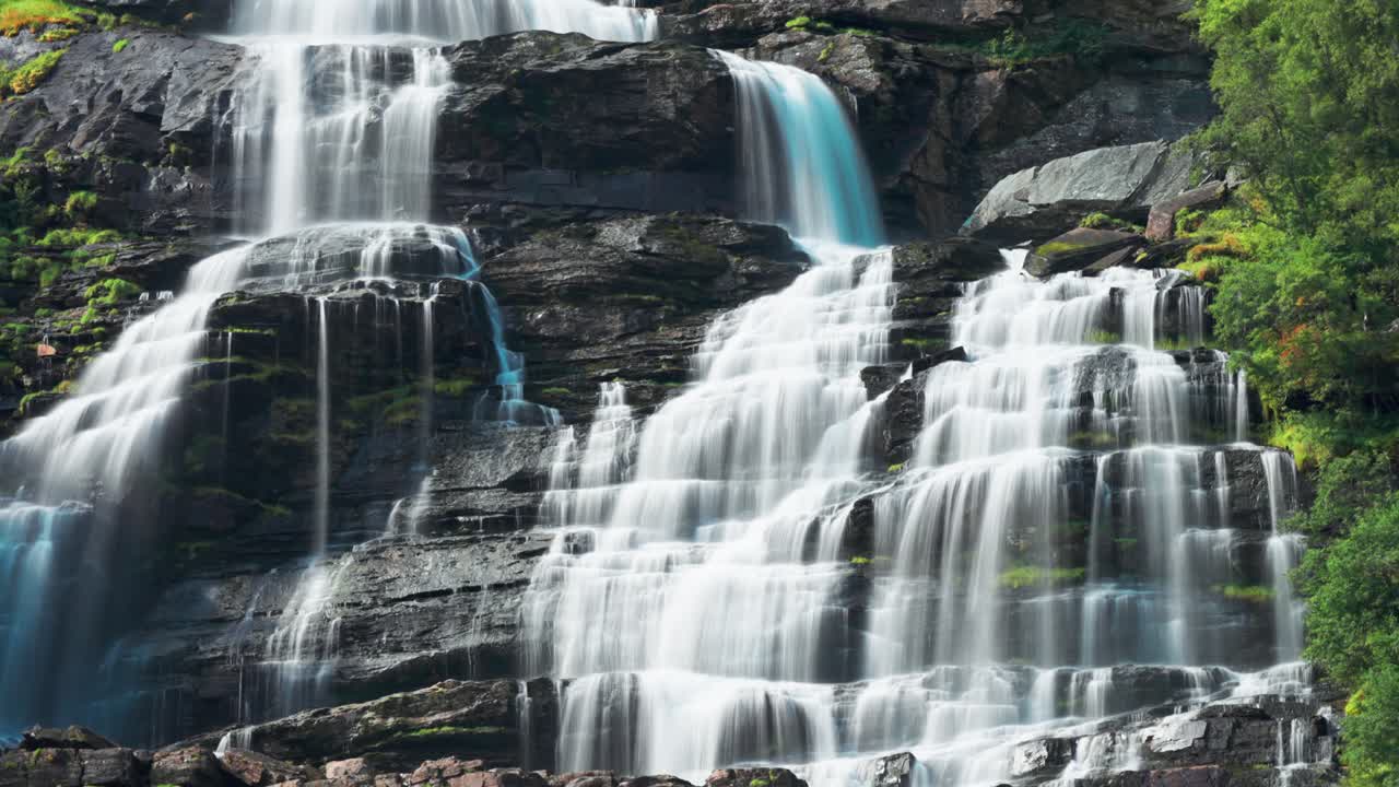 Beautiful footage of a waterfall flowing over a rocky precipice, surrounded by dense forest and bathed in natural light, creating a serene atmosphere