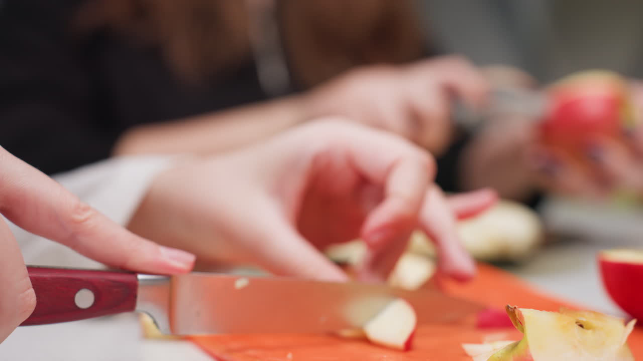 Hand view youth slicing apple pieces with kitchen knife on orange plastic cutting board while friend scrapes apple skin in background, kitchen teamwork, fresh fruit prep, clean home cooking scene