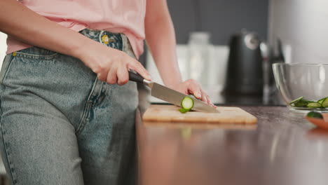 partial view of lady slicing cucumber on wooden board beside transparent glass bowl filled with sliced pieces while kitchen background appears softly blurred with calm lighting