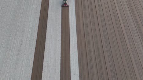 Tractor fitted with a leveling harrow moves beneath the drone in slow motion, smoothing freshly tilled earth into uniform dark brown furrows alongside pale loam strips across a vast agricultural field