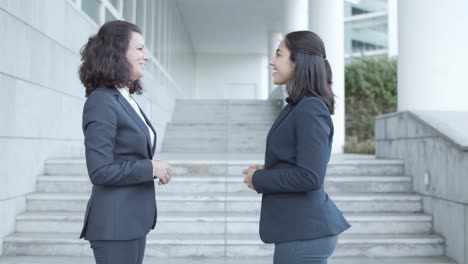 side view of two friendly female office colleagues in suits meeting outside, shaking hands and talking