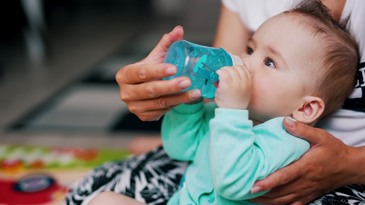 Thirsty little beautiful kid drinking water from a bottle. Caring mom supporting her child and bottle for him. Close up.