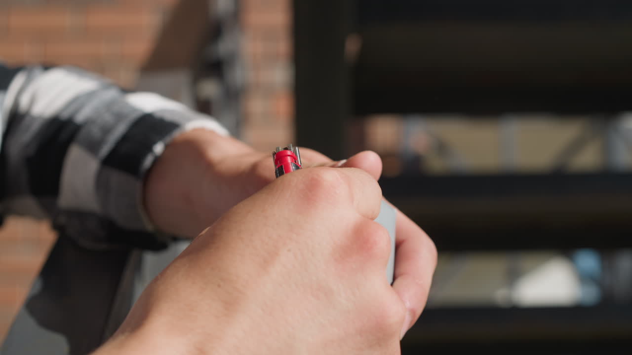 Close up of smoker retrieving cigarette from pack while holding lighter and preparing to light it, with cigarette approaching mouth in dramatic sunlight and shadows over urban staircase background