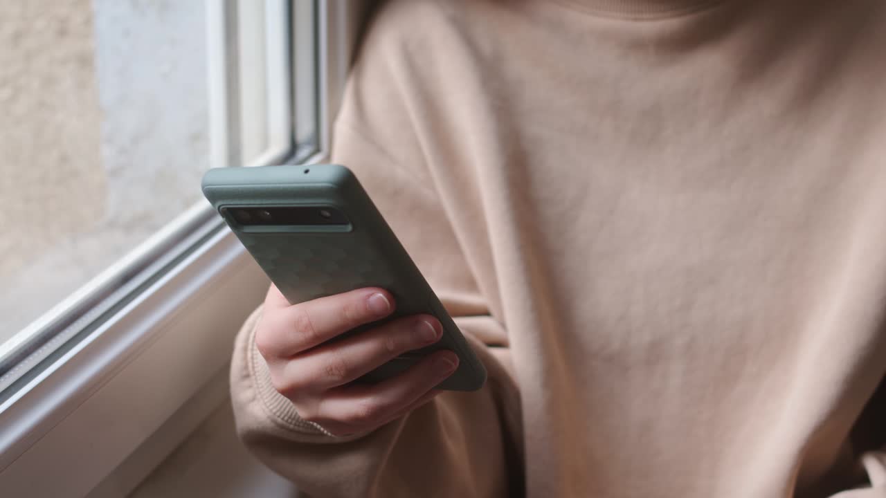 A Person in a Sweater is Using Her Mobile Phone by the Window - Close Up