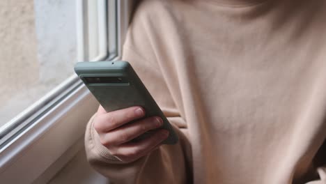a person in a sweater is using her mobile phone by the window - close up