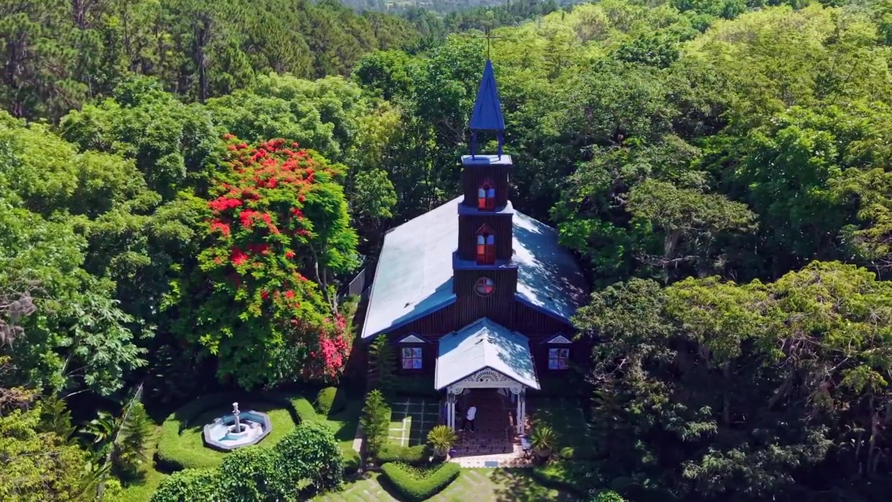 La Capilla La Milagrosa se alza con paredes de madera oscura y un campanario azul en medio de un bosque tropical en San José de las Matas, República Dominicana, con árboles en flor cerca de la entrada tranquila. Vista aérea con dron