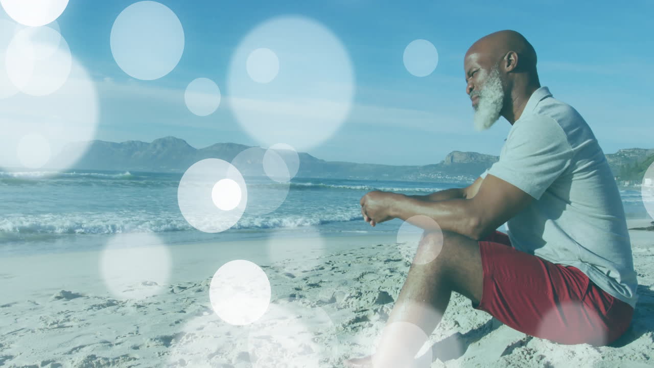 Senior man sitting on sandy beach, with animated bokeh lights illustrating health meditation