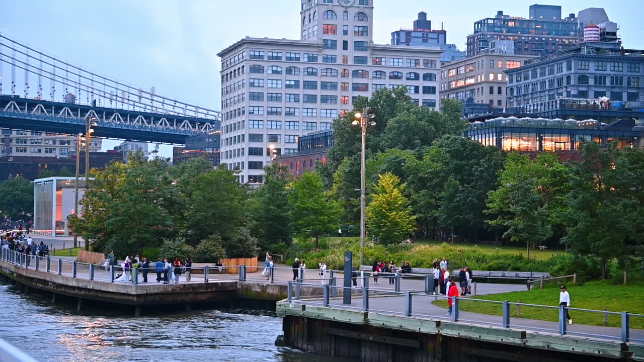 Lots of people walk by the quay with a park. High-rises of New York and the Manhattan Bridge at backdrop