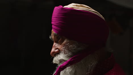 close up low key studio lighting shot of senior sikh man with beard tying fabric for turban against dark background