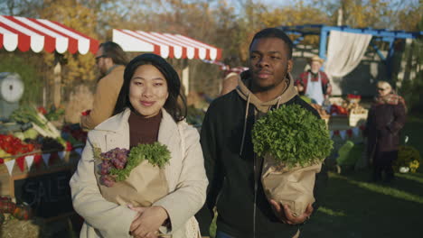 diverse couple after shopping at farmers market diverse couple standing with paper bags of vegetables or fruits spouses looking at camera smiling people buy healthy and fresh food on background shopping at local farmers market portrait view