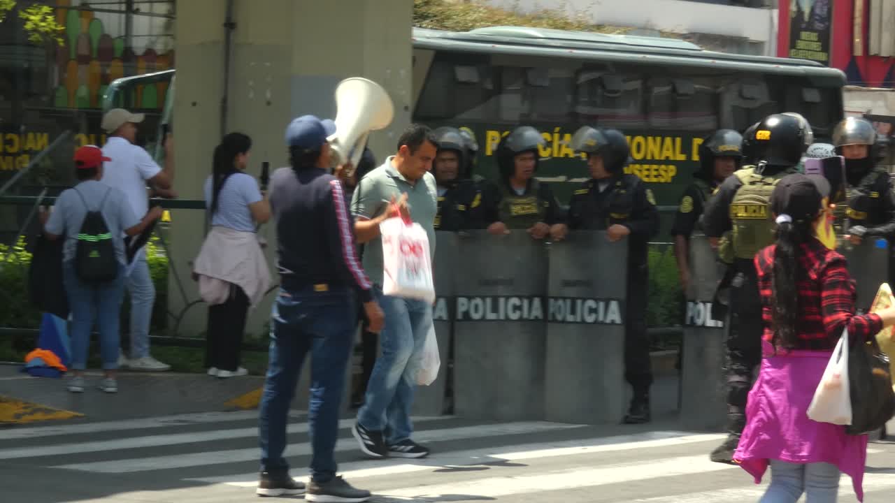 A small protest against APEC 2024 summit in Lima, Peru. A man stands with a megaphone and a lady banging on a pan in front of a line of policemen with riot shields.
