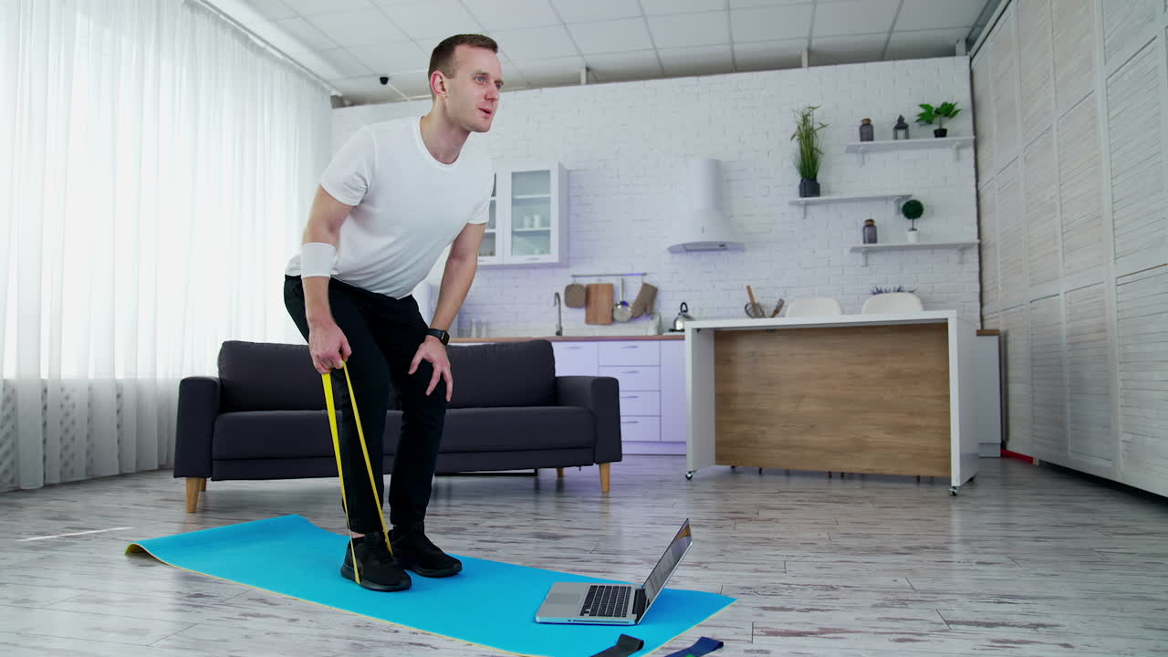 Young man training at home with a sport rubber band. Guy exercising on a mat standing in front of a laptop in kitchen. Healthy lifestyle.