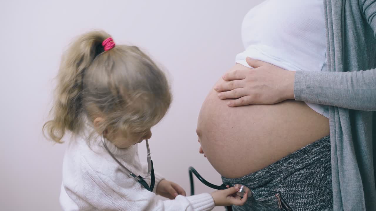 una ragazza con uno stetoscopio tocca la pancia della mamma ascoltando il bambino