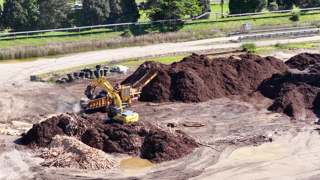 Drone captures an excavator moving soil and mulch in a sunny, industrial landscape with greenery in the background