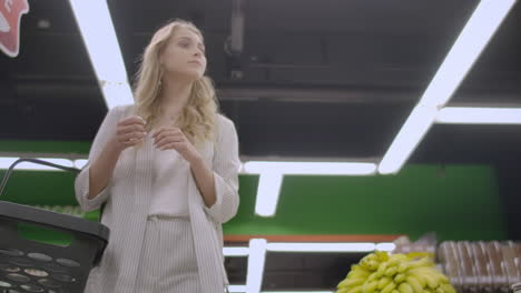 pretty lady in casual clothes is walking in grocery store steering shopping trolley with food inside it and looking around at shelves with products. women and shops concept.