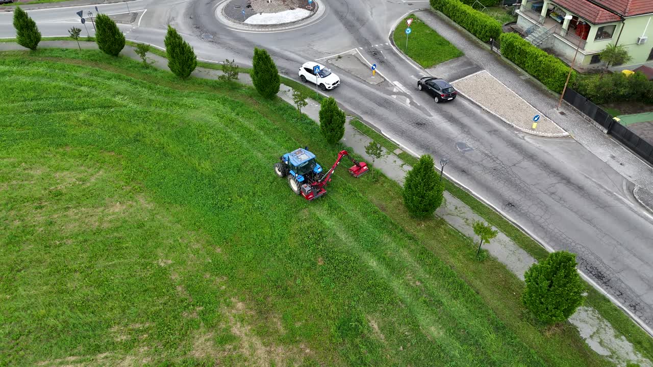 Blue tractor cutting grass in a residential area of Piacenza, Italy, near a roundabout, offering an aerial perspective of urban landscaping